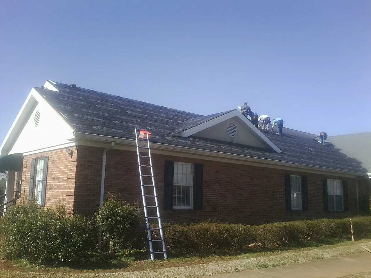 Crew tearing shingles off a brick ranch roof with a ladder set — Emergency Roof Repair work by our crew in Locust Fork