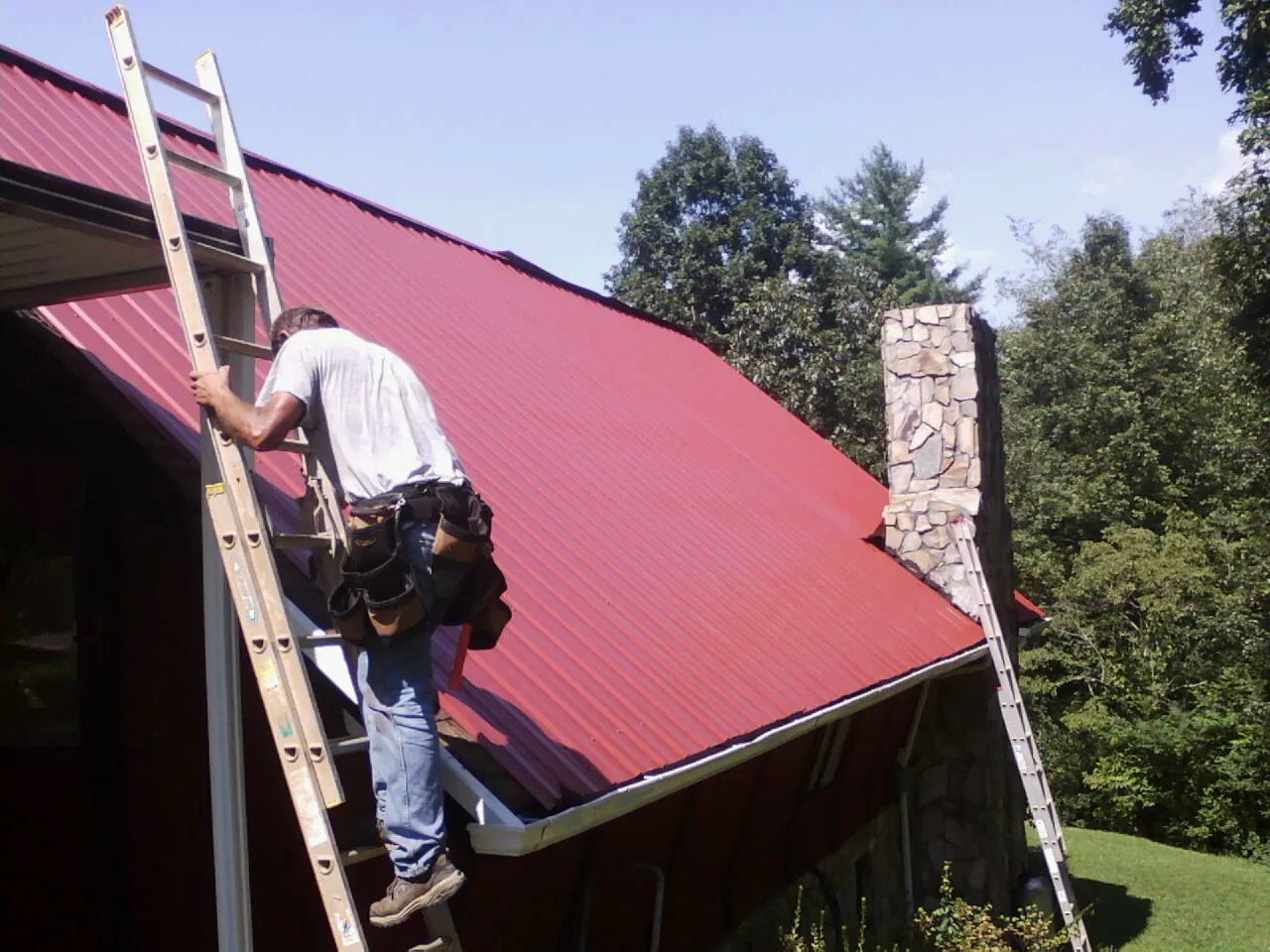 Roofer on a ladder beside a red metal roof and stone chimney — Soffit & Fascia Repair by Roofing Brothers in Locust Fork